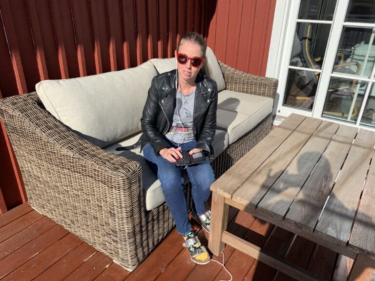Anna Bergholts sitter i en rottingsoffa med sin mobiltelefon i knäet ute på sin altan. Hon bär solglasögon med röda bågar, svart skinnjacka, blå jeans och sitt mörkblonda hår uppsatt i hästsvans.