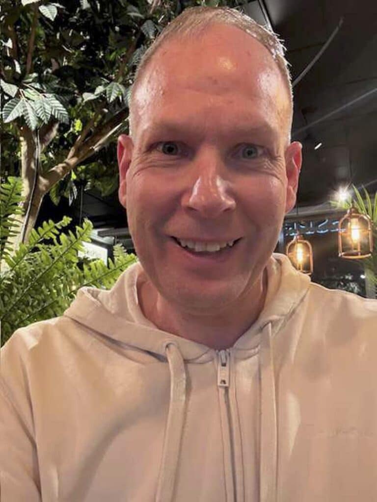 Martin Persson porträttfoto. Han har kort blont hår, ler brett, klädd i en vit munkjacka. I bakgrunden syns dämpad belysning och gröna växter.