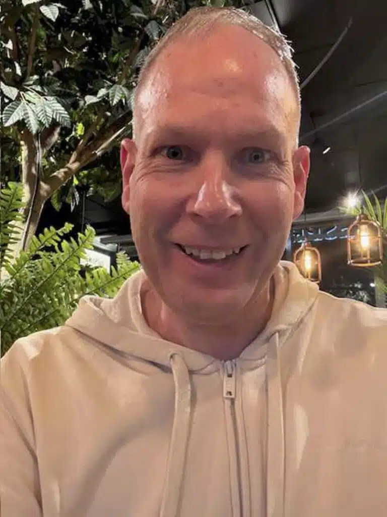 Martin Persson porträttfoto. Han har kort blont hår, ler brett, klädd i en vit munkjacka. I bakgrunden syns dämpad belysning och gröna växter.