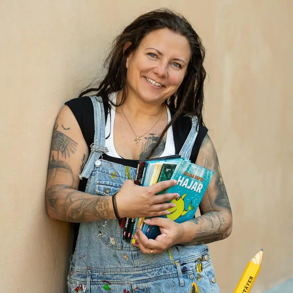 Porträttbild av Emmaill där hon håller i sina barnböcker. Hon har mörkbrunt långt hår, tatueringar på armarna och är klädd i svart topp under ljusblå hängselbyxor.