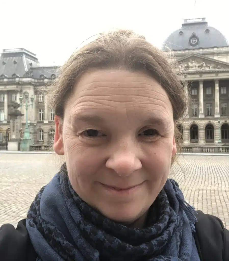 Jenny Johansson. Hon brunt långt hår och ler. Hon har på sig en blåsvart halsduk och en svart överdel på klädseln. I bakgrunden syns ett torg med kullersten.