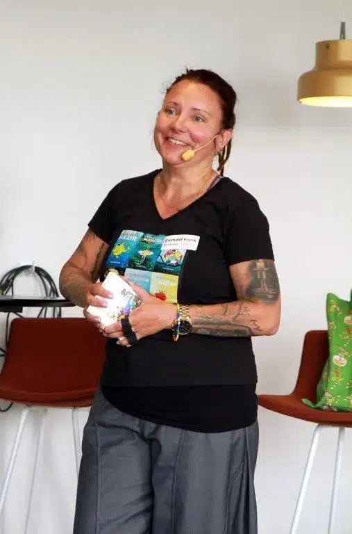 Emmalill föreläser med ett gult headset. Hon har mörkbrunt hår uppknutet i hästsvans och ler mot publiken medan hon håller i en bok i handen. Hon är klädd i svart t-shirt med olika illustrationer på framsidan, gråa byxor och ett färgglatt armband på ena handleden. I bakgrunden syns konferensstolar och en vit vägg.