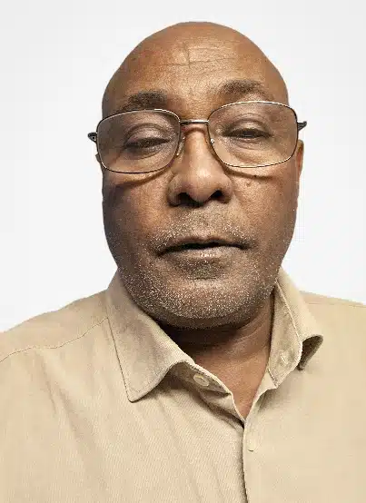 Profilbild Yakem. Han har mörk hy, glasögon, rakat huvud, beige skjorta.