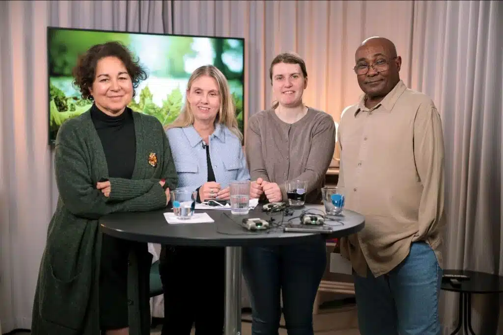 Sabina, Anna, Sandra och Yakem står tillsammans vid ett runt bord med draperier och en tv-skärm bakom sig. Sabina har mörkt lockigt hår, grön lång stickad rock, Anna med långt ljust hår har en ljusblå kavaj, Sandra har mörkt brunt hår klädd i en ljusgrå tröja, Yakem har glasögon, rakat huvud, beige skjorta och blå jeans.