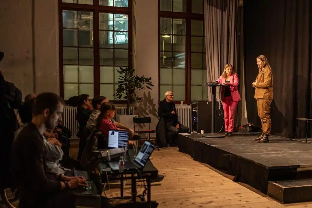 Anna Bergholtz i rosa kavaj och Susanna Cederquist i ljusbrun kavaj står på scen framför publik. Samtalet textas live på overheadduk av några personer som sitter vid datorer nedanför scenen.