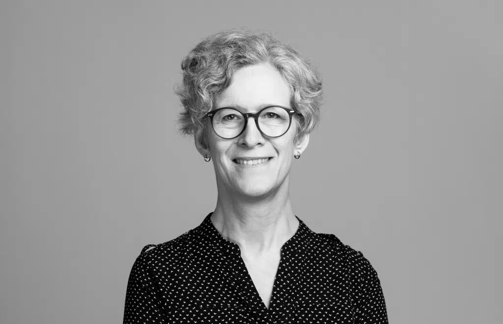 Sara Rydin porträttfoto i svartvitt. Sara har kort lockigt hår, mörka rundade glasögon och har på sig en mörk överdel med prickigt mönster.