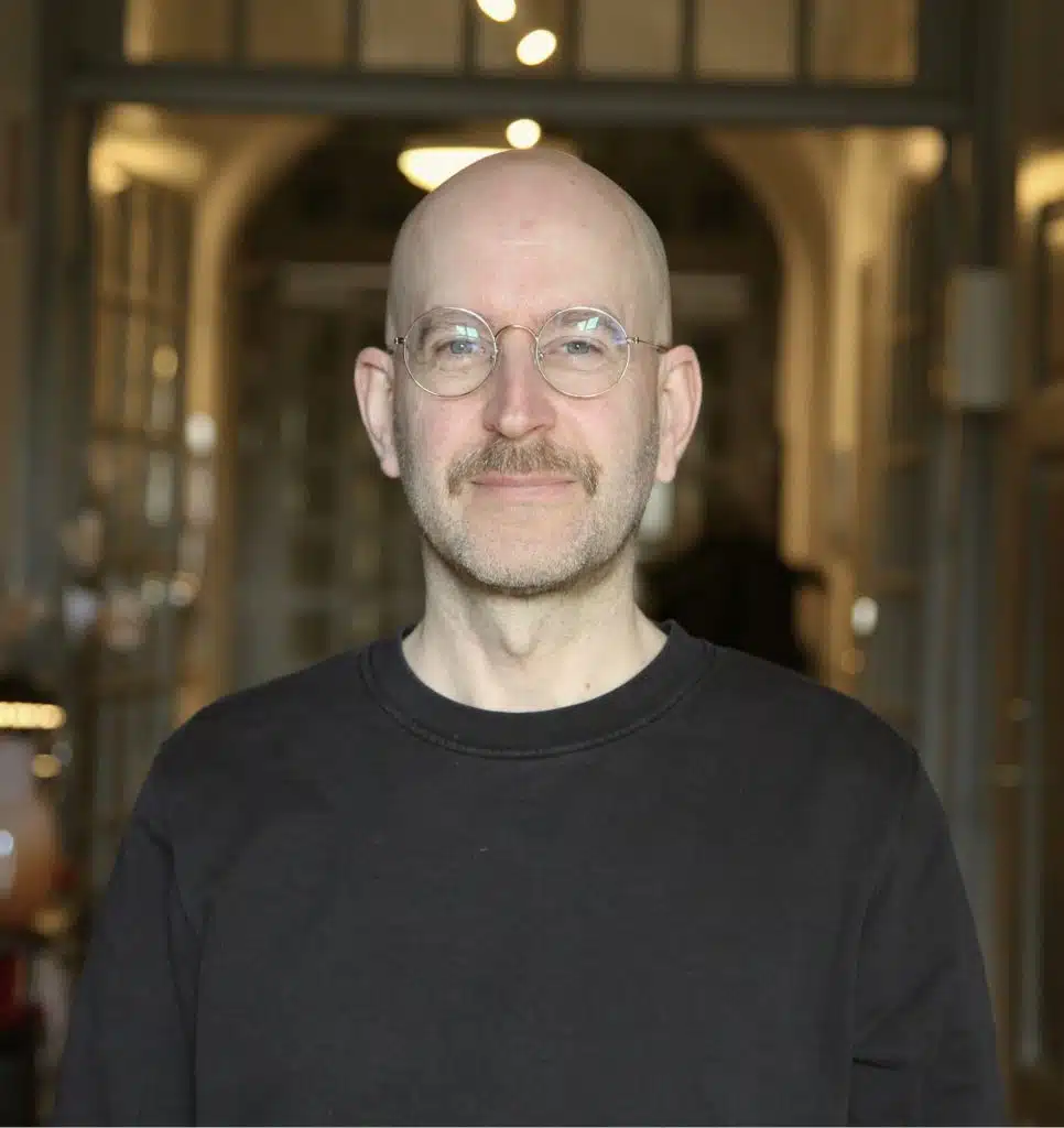 Porträttfoto Gunnar Bark. Han har runda glasögon, rakad huvud, klädd i en svart tröja och står i en lokal med dämpad belysning.