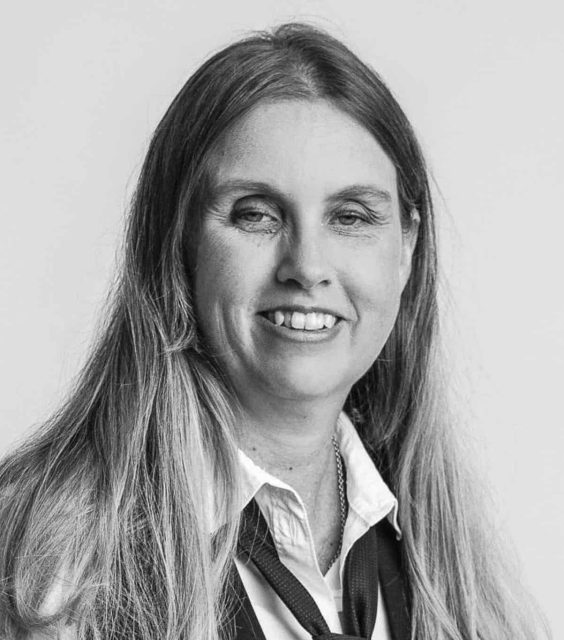 Svartvit porträttbild av Anna Bergholtz. Hon har långt blont hår och ler. Hon har på sig vit skjorta, svart slips och väst.