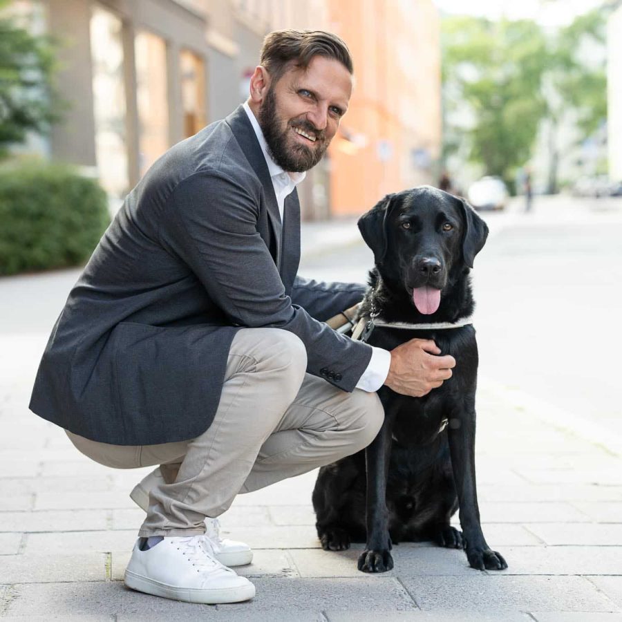 Fatmir Seremeti står på knä intill sin svarta ledarhund på en gata. Fatmir har brunt kort hår, skägg och mustasch. Han är klädd i grå kavaj, vit skjorta, ljusa byxor och skor.