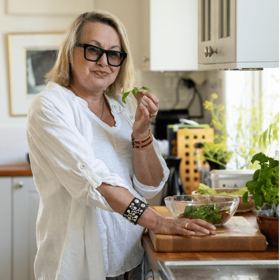 Louise Hoffsten står i ett kök och håller i ett grönsaksblad. Hon har axellångt blont hår, svarta tjocka glasögon, vit skjorta och beigefärgade byxor. Framför sig på köksbänken syns en genomskinlig skål med grönsaker.